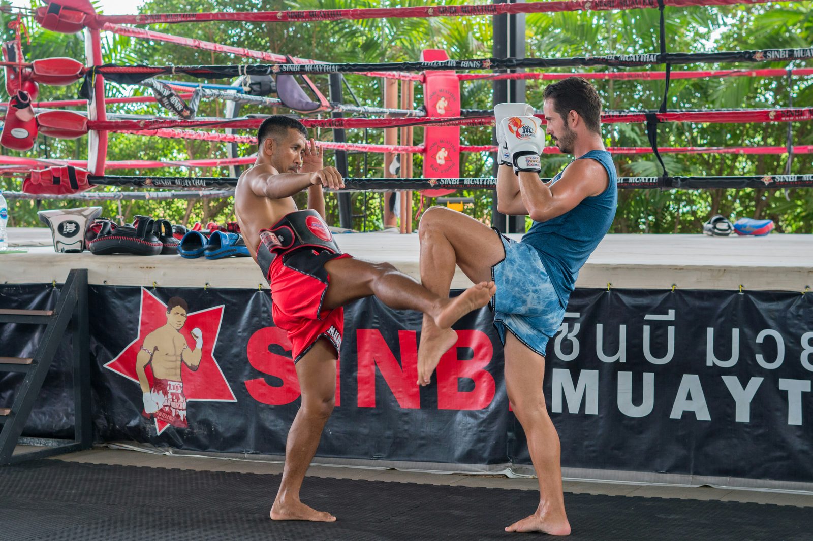 Two training partners practice Muay Thai beside the Sinbi ring, with red ropes and green foliage behind them.